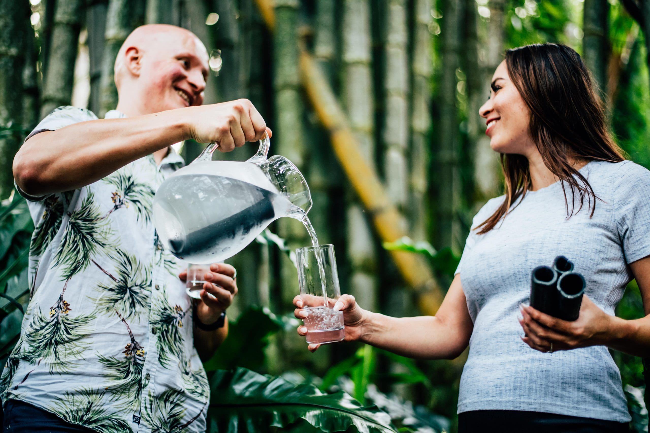 A man in a tropical shirt pouring water from a jug into a glass held by a smiling woman in an outdoor setting with lush green bamboo in the background.